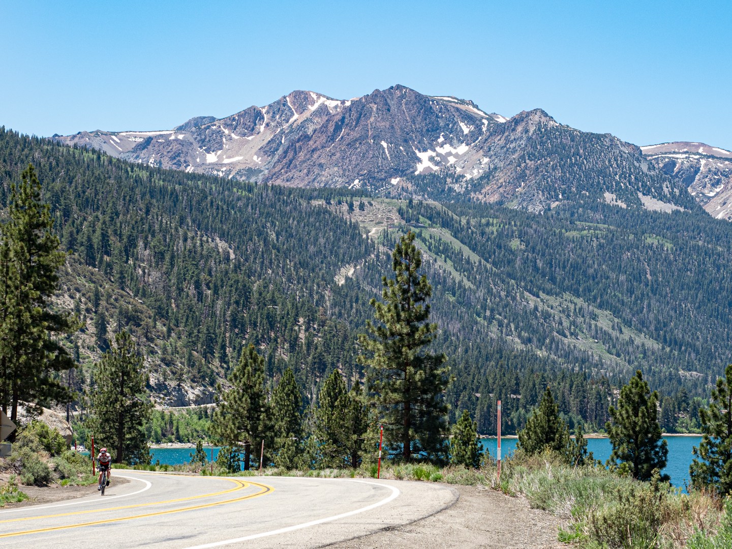 cyclist on shoulder pedaling up June Lake Overlook with the mountains and June Lake behind him